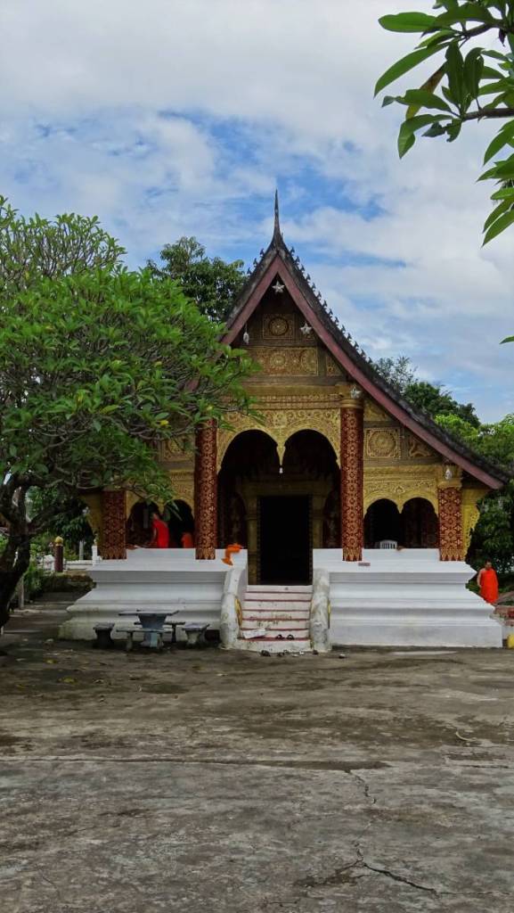 A small Buddhist temple in Luang Prabang with a sloped roof, golden facade and red pillars. A tree grows in front while some monks i orange robes sit on the porch.