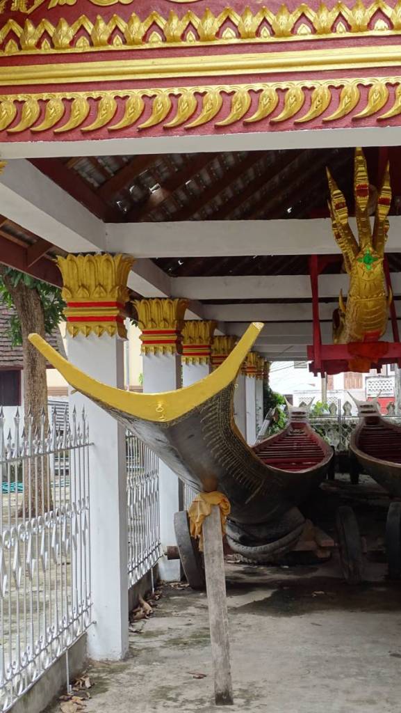 Two long, narrow, wooden boats used for boat racing festival and a golden statue of a 3-headed snake-dragon stored under the roof at the temple compound in Luang Prabang.