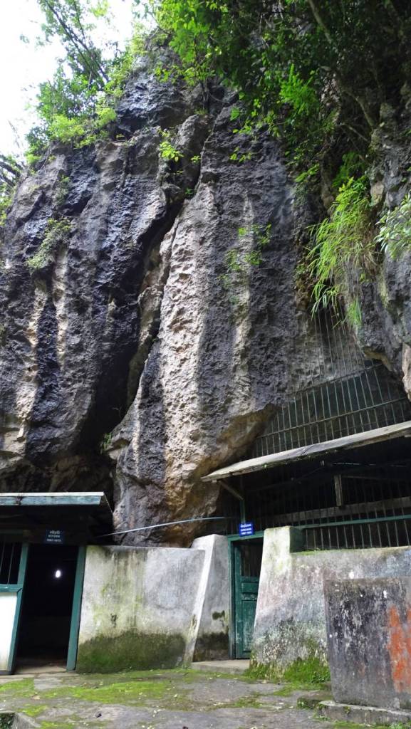 The entrance to the caves in Vieng Xai with a moss covered concrete wall, wooden doors and a crate over the large opening in the rock.