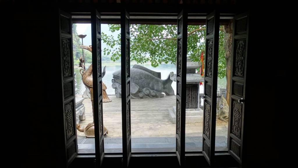 A view at the brass crane and stone fish statues through the wooden doors of Thanh Cao Son pagoda in the scenic Trang An area