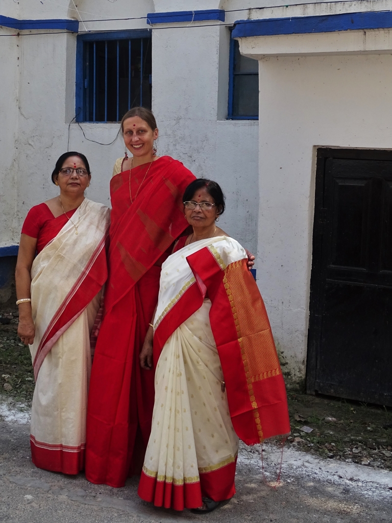 Weronika stands holds in her arms her mother in law and Sayak's aunt, all of them dressed in saris for the ocassion of Durga Puja