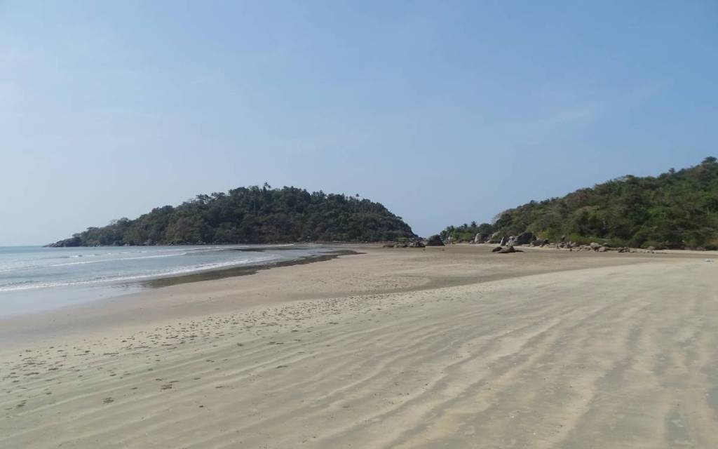 Empty stretch of sand at the very tip of Palolem beach with a scattering of boulders and a forested Cancona Island