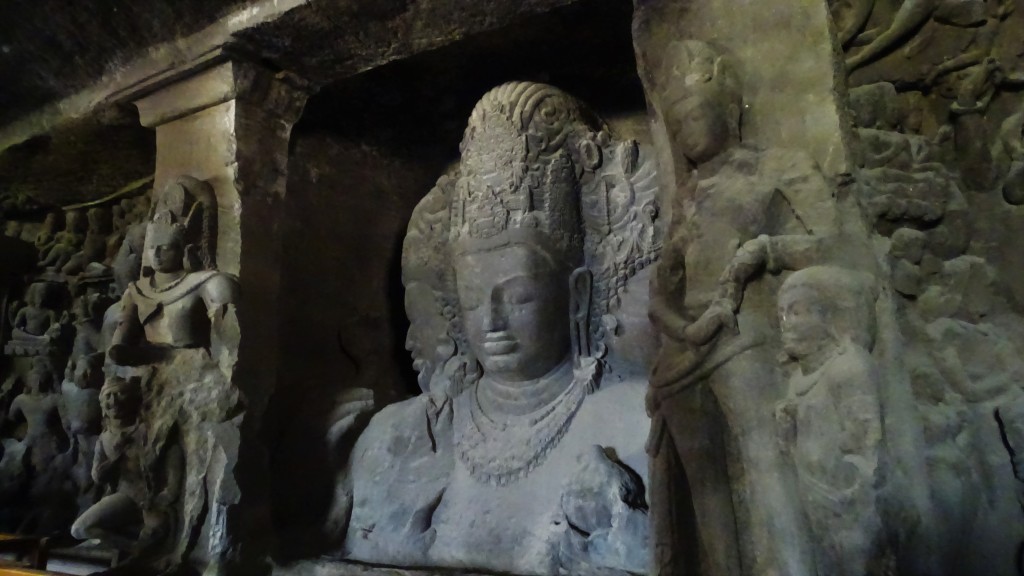 A stone sculpture of a three-headed God, surrouned by many smaller statues of Hindu gods inside the Elefanta Cave