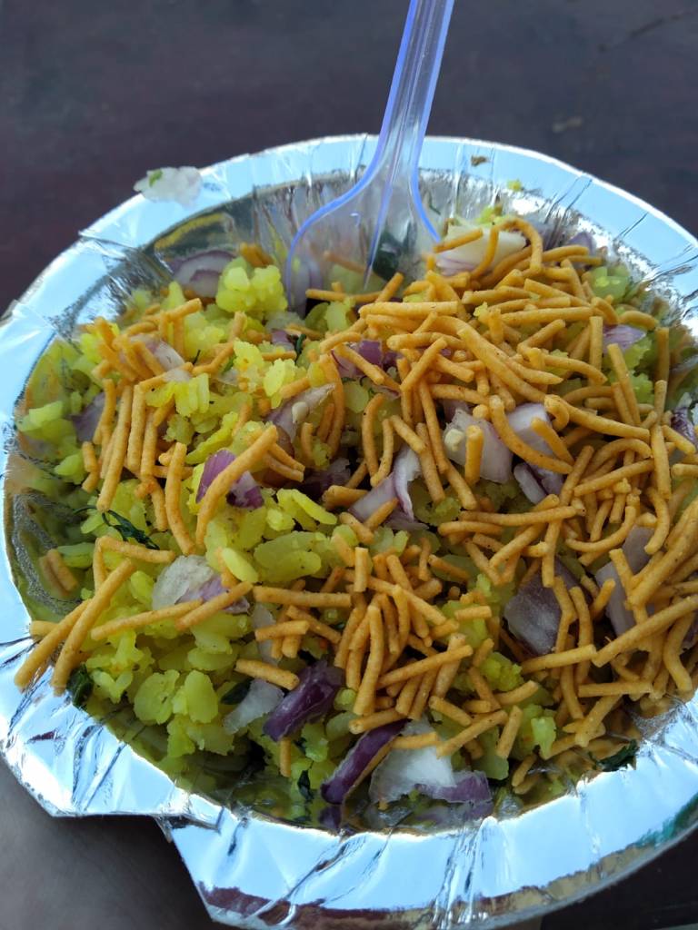 an aluminium foil bowl filled with poha: yellow flattened rice, onion and coriander 